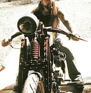 A photo of a long haired blonde male riding his oldschool Harley motorcycle with jeans and a shirt